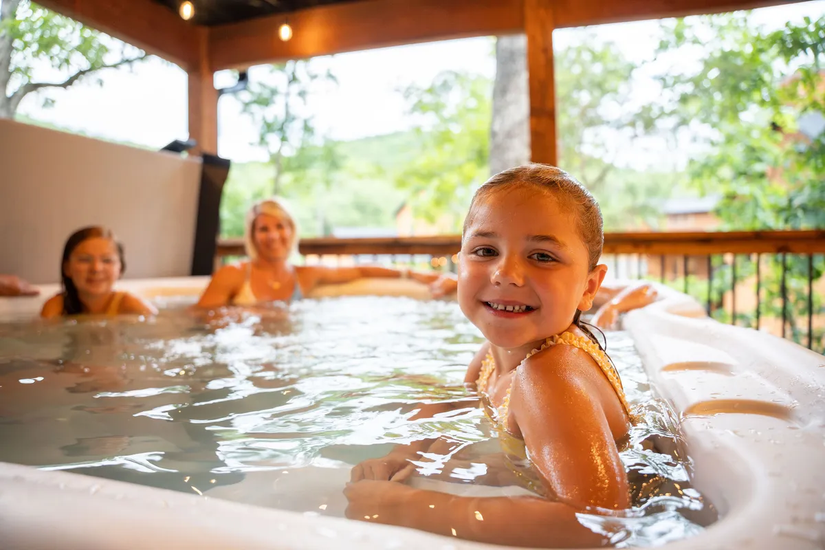 A hot tub nestled indoors, surrounded by lush greenery visible through the windows. A smiling child relaxes in the tub, enjoying a moment of family bonding and tranquility while being close to nature.