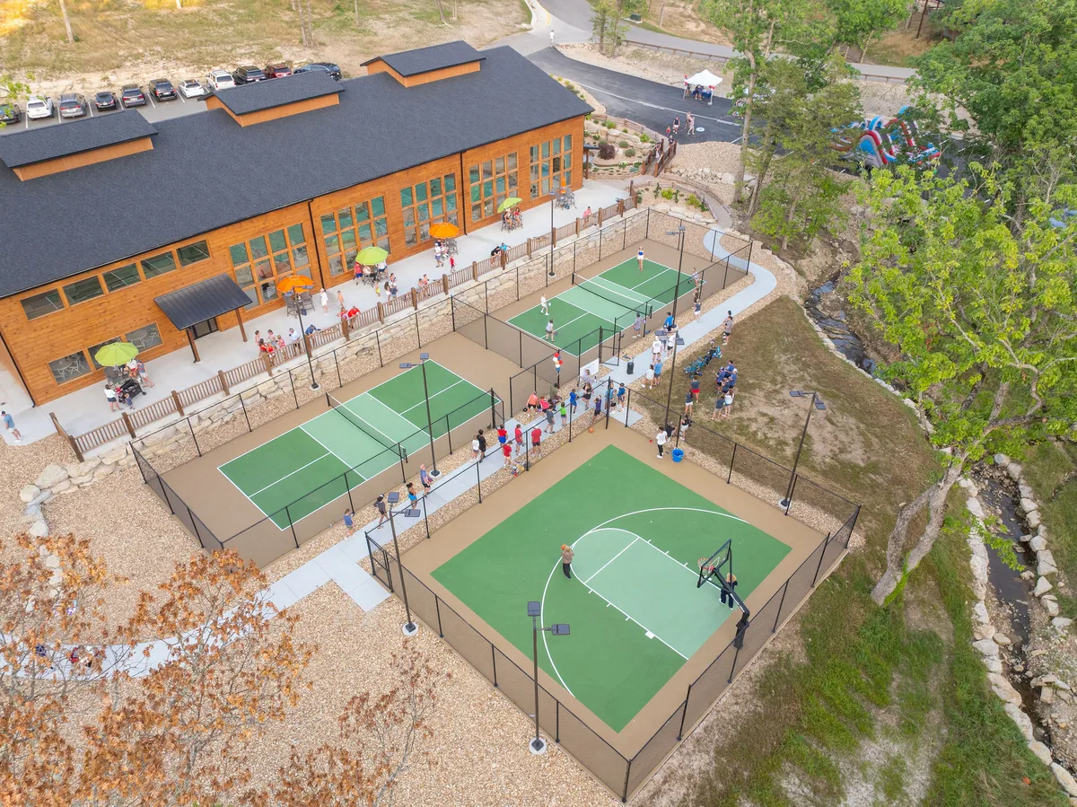 A dynamic exterior showing sports courts in use, including people actively playing basketball and tennis. The recreational facility provides a backdrop, with other people gathered and engaging in outdoor play.