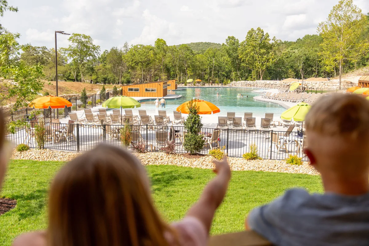 A vibrant outdoor swimming pool scene with colorful umbrellas and children pointing excitedly. The surrounding area is designed for gatherings, and the setting sun casts a warm, inviting glow over the pool.