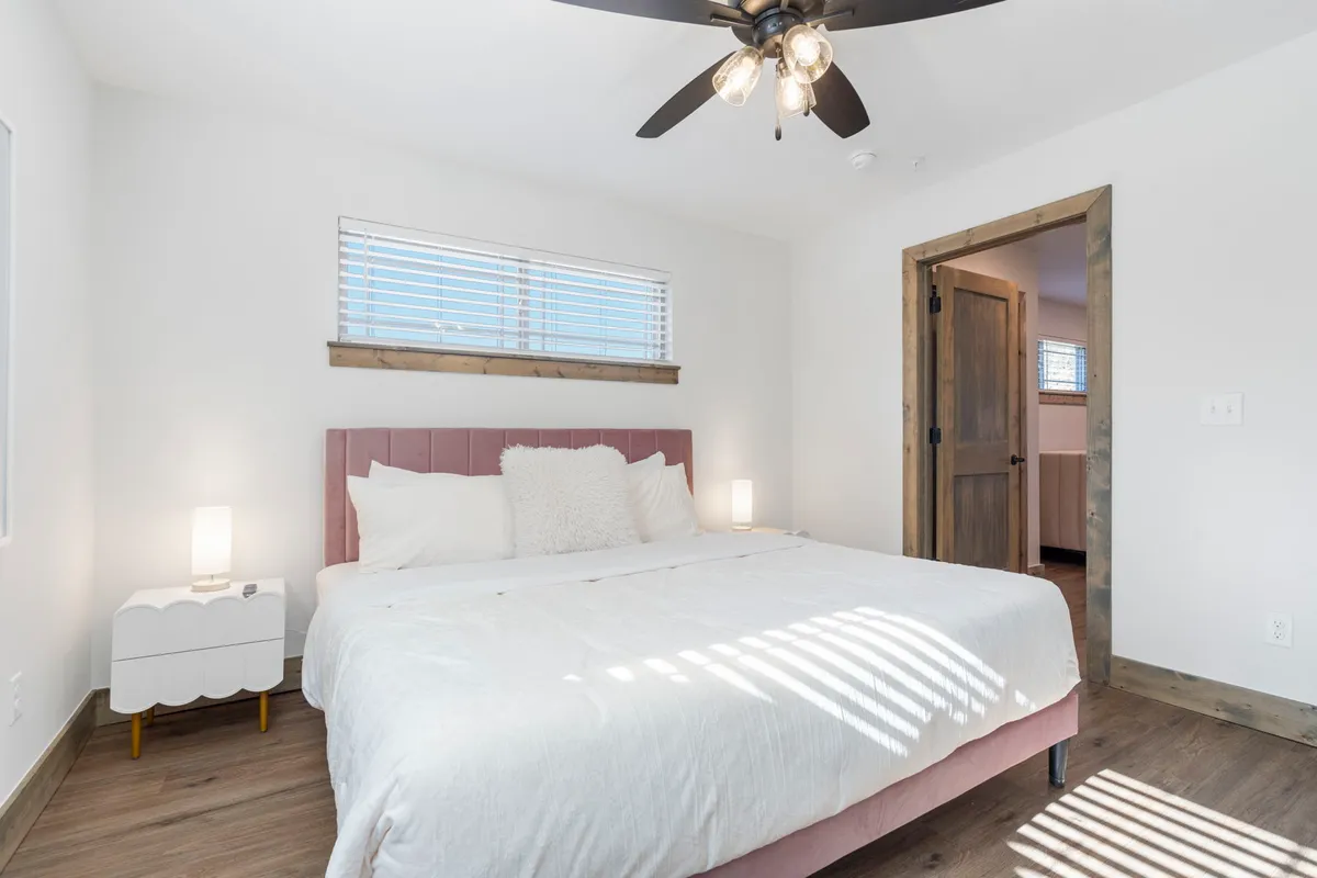A cozy, calm king-size bedroom featuring a generously sized bed with decorative pillows, a nightstand with a lamp, and a wooden door. Soft, natural light filters into the room, highlighting the restful decor and inviting bedding.