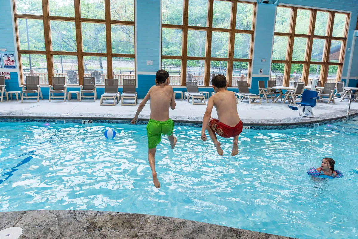 An indoor swimming pool where two boys are jumping into the water and a girl is swimming nearby. Large windows let in an abundance of natural light, emphasizing the bright and airy atmosphere. The scene captures children actively playing and enjoying time together by the pool.