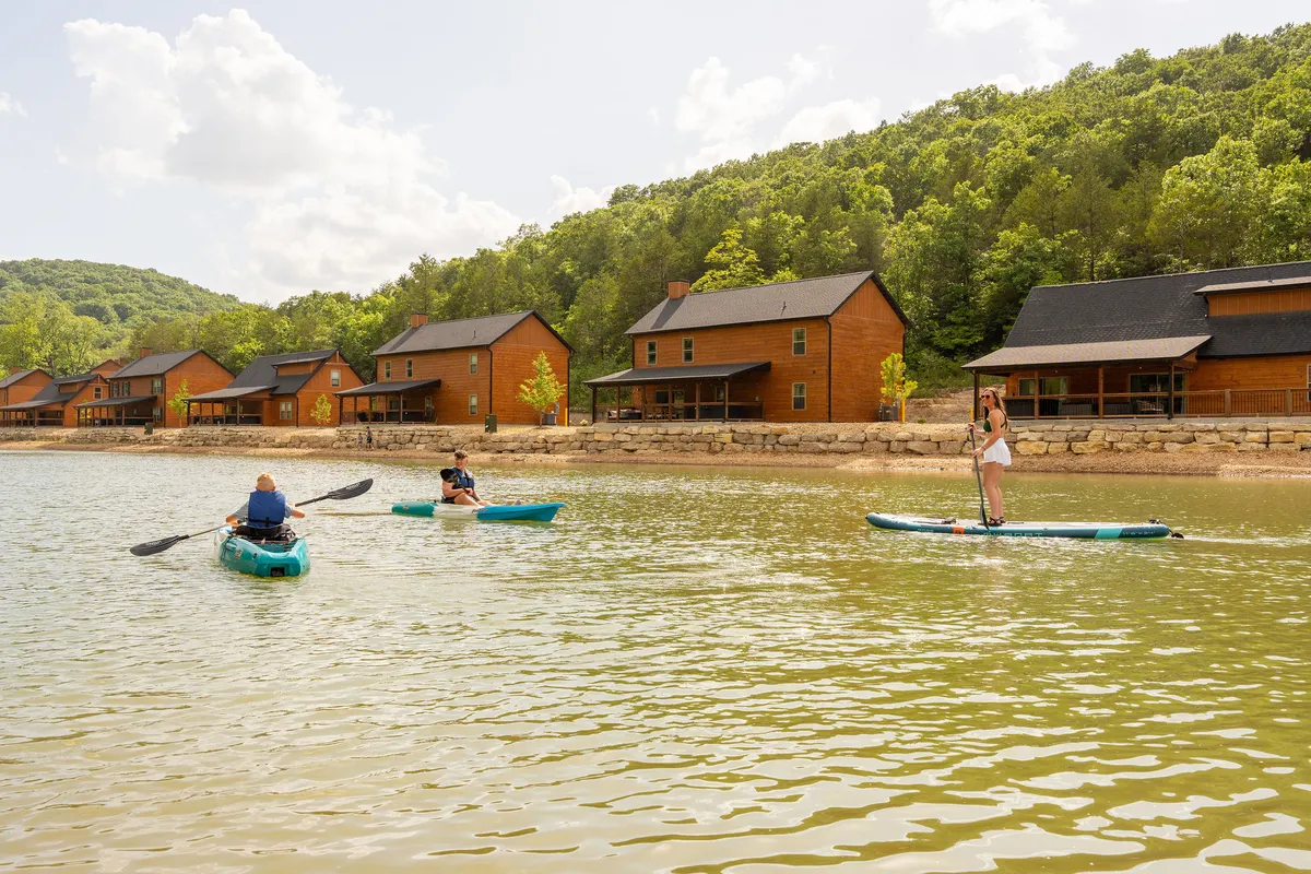 This dynamic exterior shot captures kayakers and a paddle boarder enjoying the water near a cluster of cabins, with a lush natural landscape in the background. The scene is playful and serene, reinforcing the idea of lakeside adventures, water sports, and relaxing by Table Rock Lake.